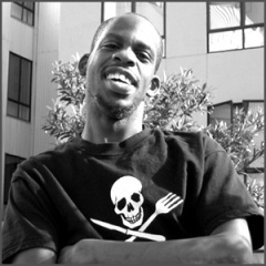 Image of Leroy Moore, smiling, with his arms crossed over his chest. He is wearing a black t-shirt with a skull and crossbones.